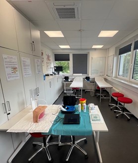 „Leerer Skills-Lab-Raum mit blauem Tuch, Infusionsbesteck, Armmodell, Monitor und Schränken auf hellem Tisch, rote Stühle.“