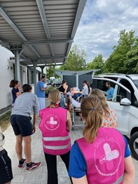 „Medizinstudenten in pinken Skills-Lab-Westen schieben eine Trage mit Patient von weißem Gebäude weg, daneben Van und Bäume.“