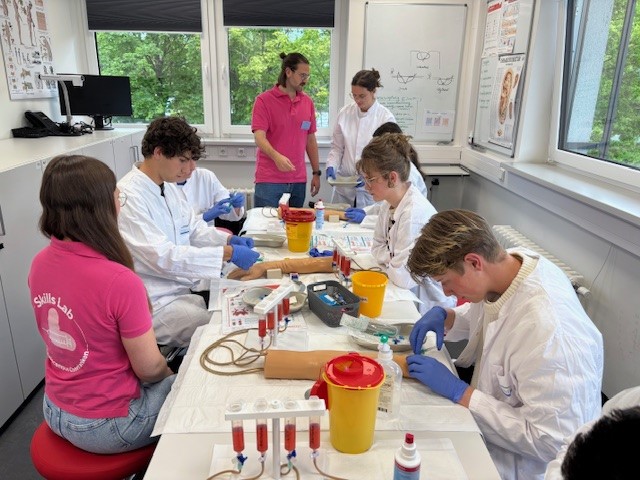 Foto, Studierende im weißen Kittel üben Blutabnahme an Modellen, zwei Tutor:innen im pinken Shirt stehen dahinter.