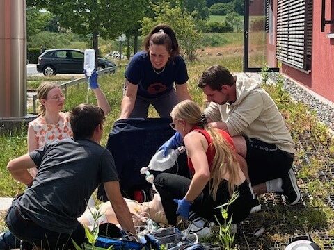 Foto, fünf Personen knien draußen um Simulationspuppe, eine Person hält Infusionsbeutel hoch. 