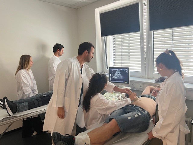 „Fünf Studierende in Kitteln beobachten Ultraschall-Simulation einer Schwangeren auf Liege in hellem Raum mit Jalousien.“