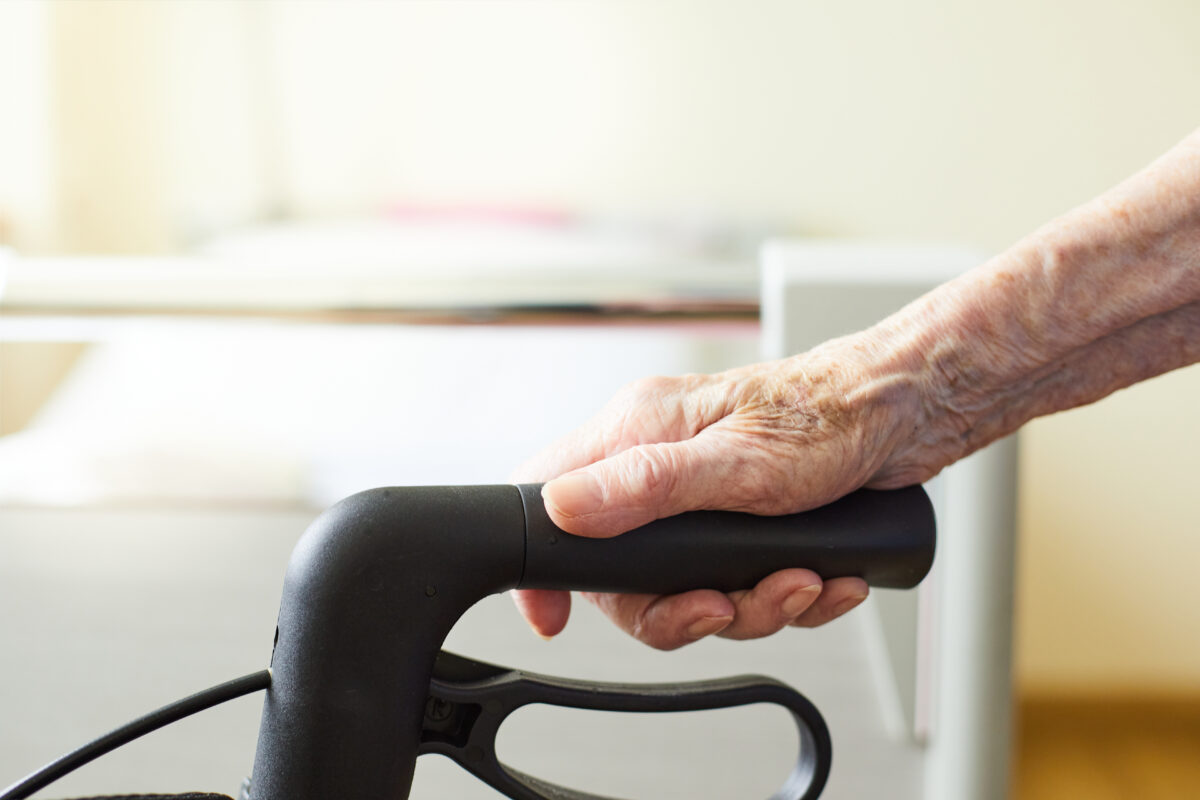 „Ältere Hand greift Rollstuhlgriff, helles Zimmer mit Tisch/Papier im Hintergrund.“