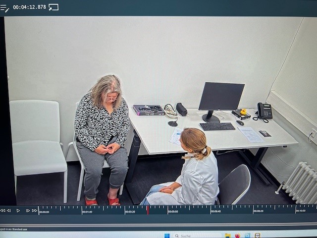 „Ärztin in weißem Kittel sitzt am Schreibtisch mit PC und spricht mit älterer Frau in karierter Bluse, weiße Stühle, Heizung, dunkler Boden.“