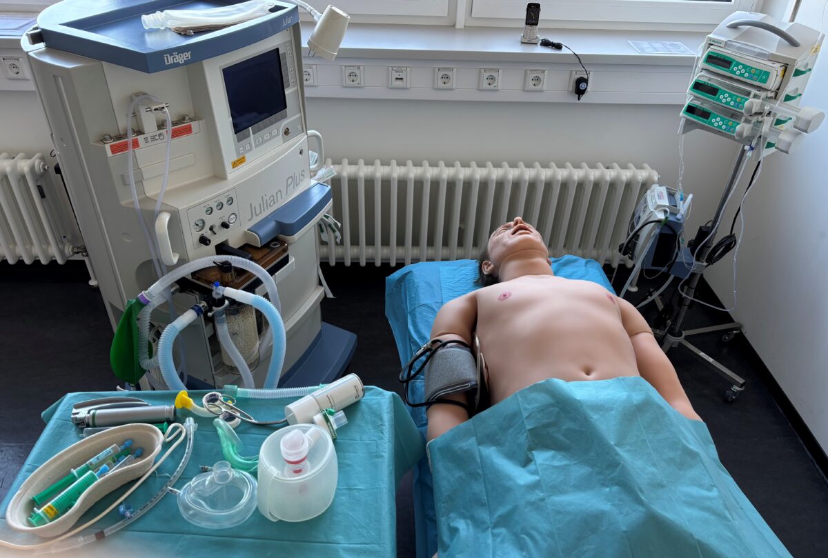 „Anästhesie-Simulation: Puppe auf Liege mit Dräger Fabius GS, Schläuche, Maske, Infusion, Heizung am Fenster.“ 
